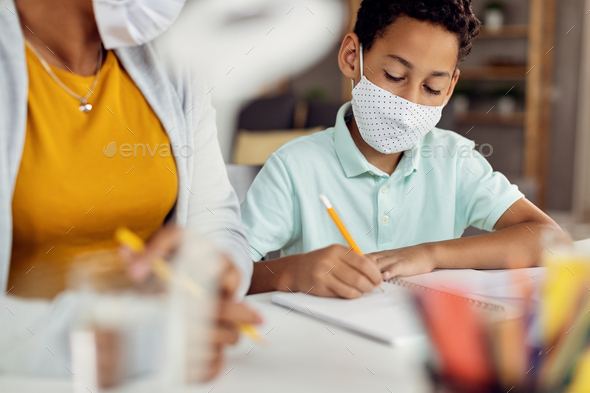 African American boy with face mask doing homework with help of his mother.
