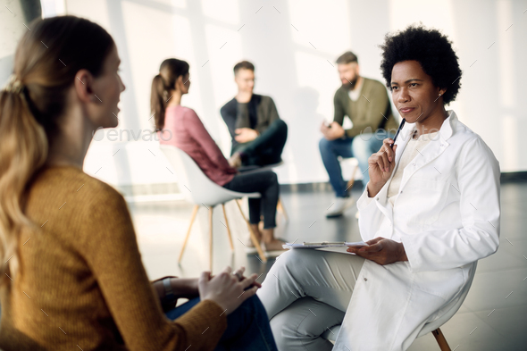 Black female doctor communicating with a woman during group psychotherapy meeting.