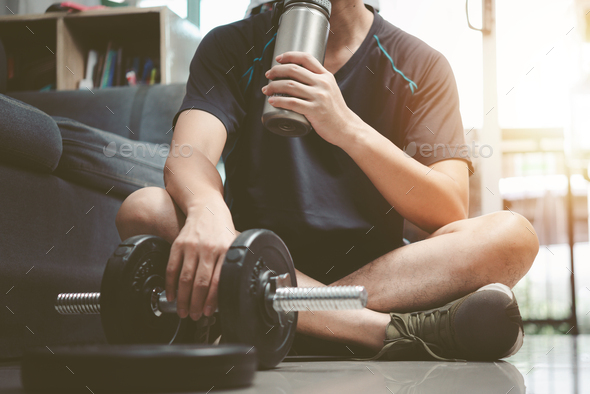 Man drinking protein or recovery drink sitting on the floor of a gym after a workout training.