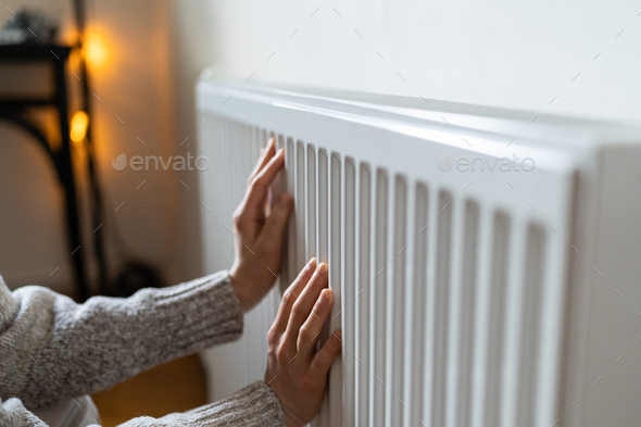 Closeup of woman in sweater puts hands on room central heating battery to warm up in cold apartment - Stock Photo - Images