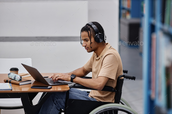 Black teenage boy with disability studying in college library and using laptop - Stock Photo - Images