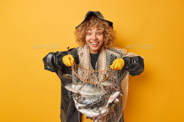 Horizontal shot of positive curly haired female fisher holds net full of fish boasts of her caught h