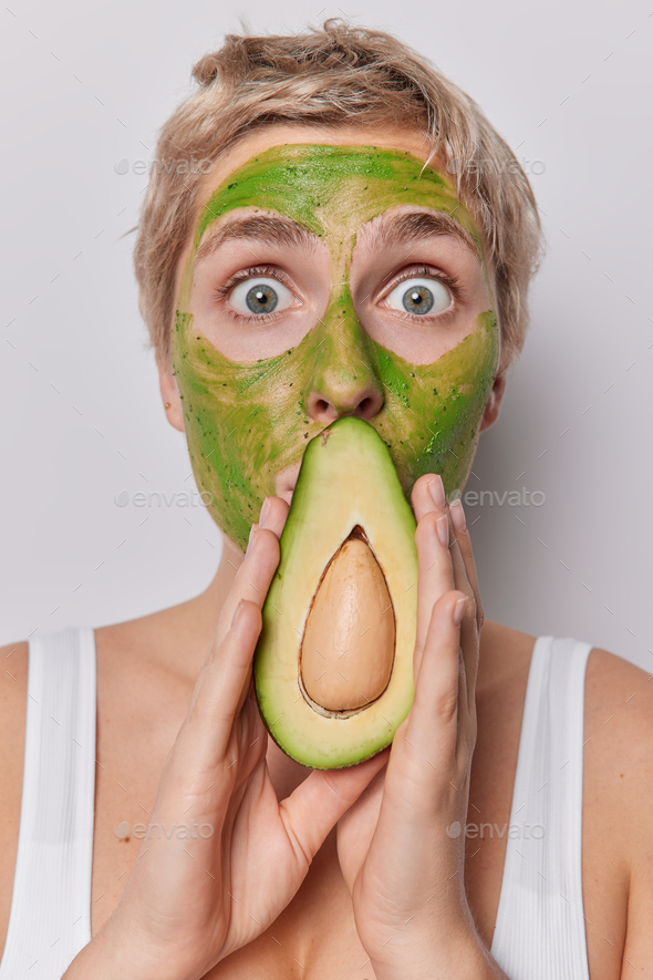 Close up portrait of surprised blue eyed woman with short hair covers mouth with half of avocado app