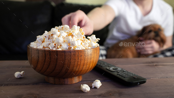 Wooden bowl with salted popcorn and TV remote on wooden table.