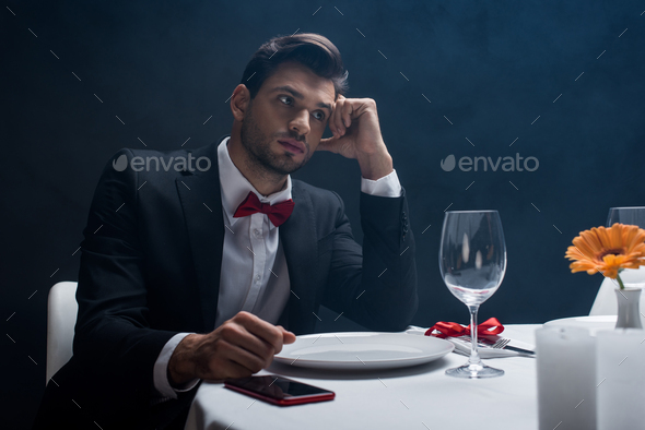 Elegant man with hand by head sitting by smartphone on served table on black background with smoke