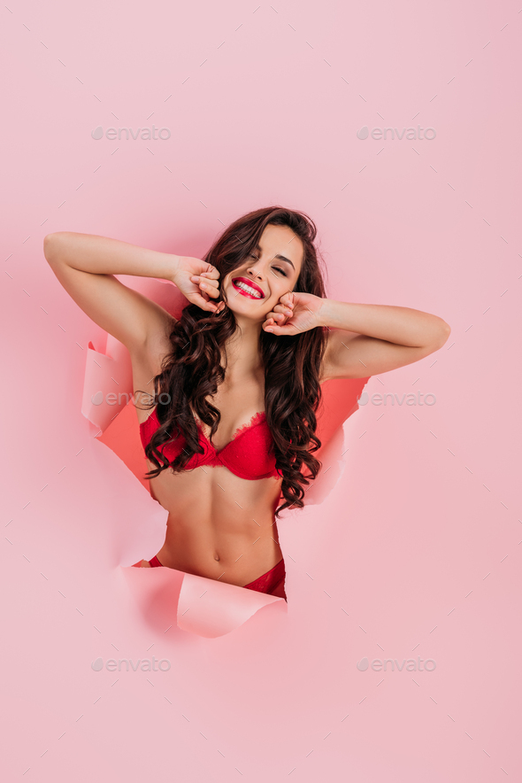 sexy, cheerful girl in red lingerie looking at camera while stretching in  paper hole on pink Stock Photo by LightFieldStudios