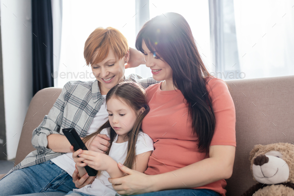 Smiling mothers sitting near daughter with remote controller on couch