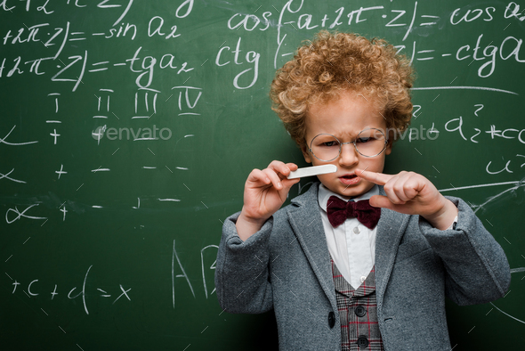 smart child in suit with bow tie pointing with finger at chalk near chalkboard with mathematical