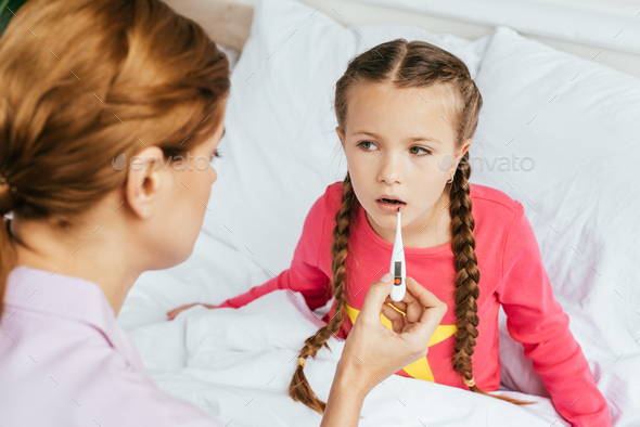 sick daughter measuring fever with thermometer while sitting in bed with mom