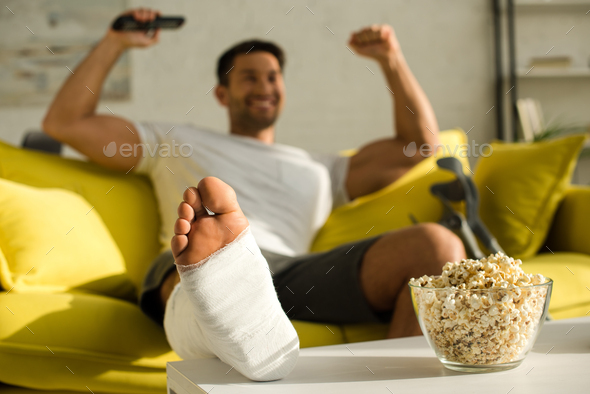 Selective focus of popcorn and broken leg of cheerful man with remote controller on couch