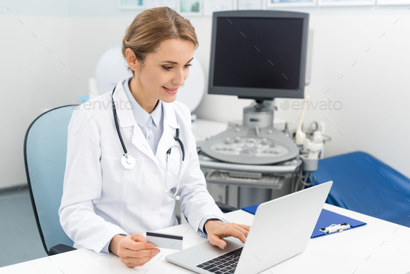 smiling doctor using laptop and credit card in clinic with ultrasound scanner
