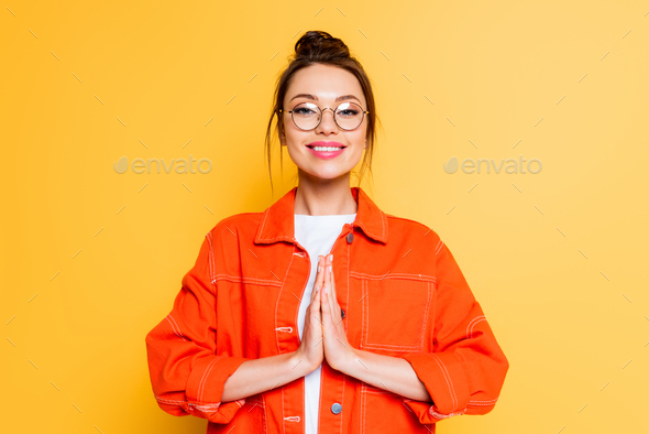 smiling student in eyeglasses standing with praying hands isolated on yellow