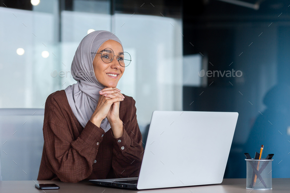 Smiling and dreamy businesswoman working inside office with laptop, woman in hijab and glasses - Stock Photo - Images