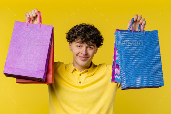 Excited man with colorful paper bags after shopping on yellow studio background. Concept of sale