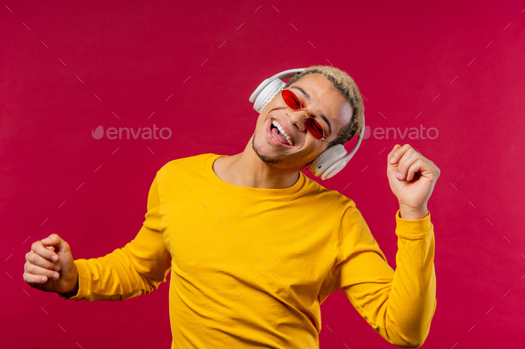 Positive american man listening music, enjoying dance with headphones on red studio background. - Stock Photo - Images
