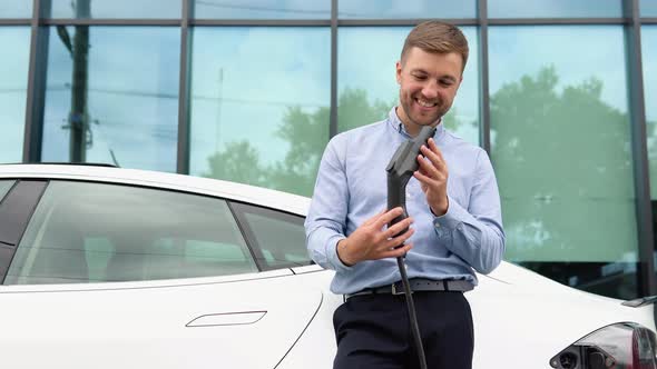 Hansome Guy Standing Near His New Modern Electric Car and Holding Plug of the Charger alt