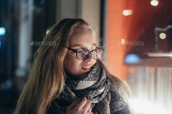 young beautiful woman in glasses on a city street at night in the light of shop windows