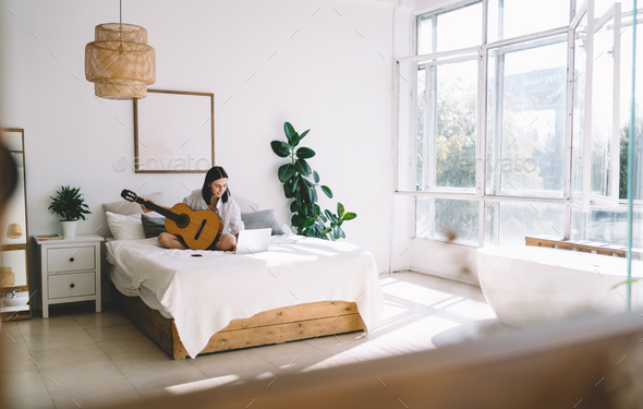 Young woman with netbook enjoying hobby with musical instrument relaxing on bed