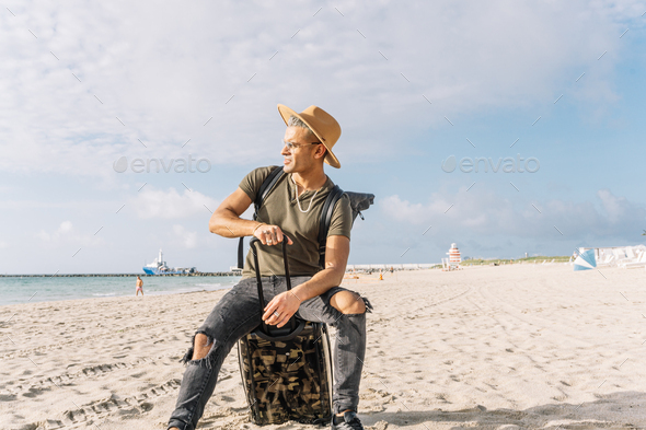 Handsome man using his carry on luggage as a chair, on the beach looking at the ocean. - Stock Photo - Images