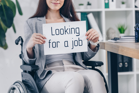Cropped view of disabled employee smiling and holding placard with looking for a job lettering on