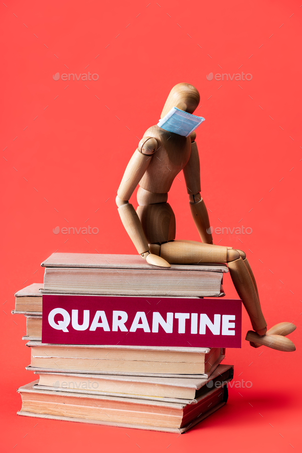 wooden doll in medical mask on books near card with quarantine lettering on red