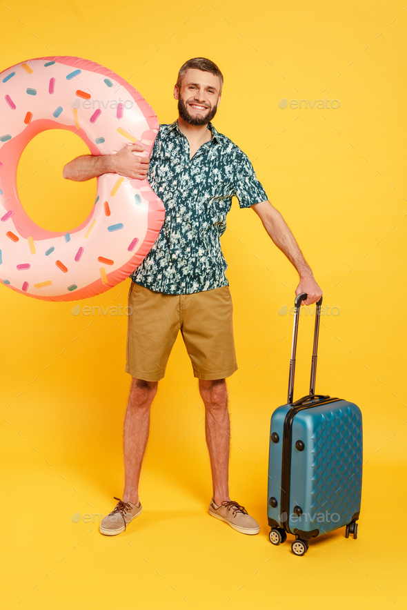 full length view of happy bearded guy with travel bag and swim ring on yellow