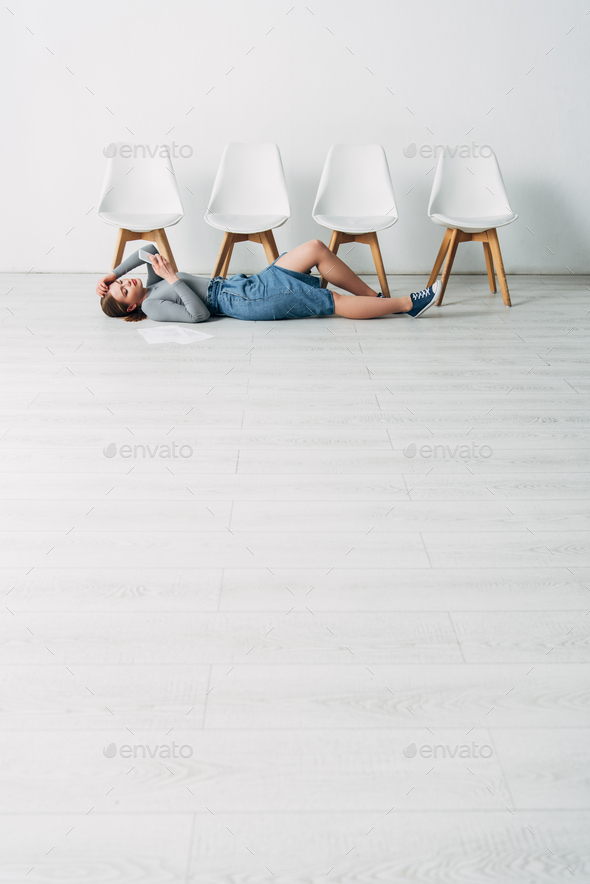 Low angle view of attractive employee using smartphone while lying near resume on floor in office