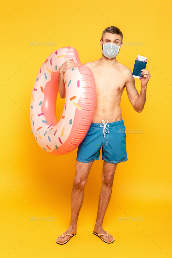 full length view of guy in swim trunks and medical mask with swim ring and passport on yellow