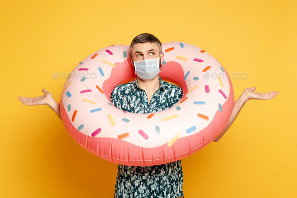 confused guy in medical mask with donut swim showing shrug gesture ring on yellow