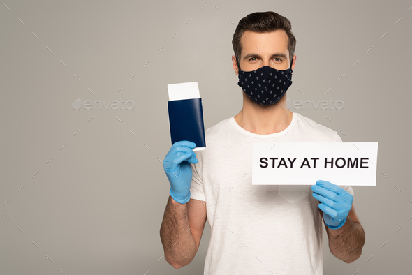 Man in safety mask and latex gloves holding card with stay at home lettering and passport with air