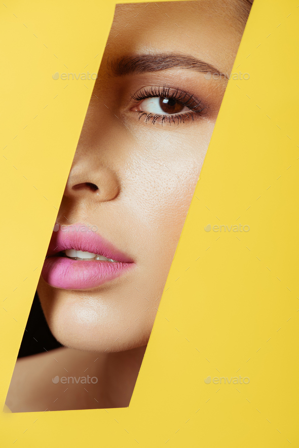 Woman with makeup looking at camera across quadrangular hole in yellow paper