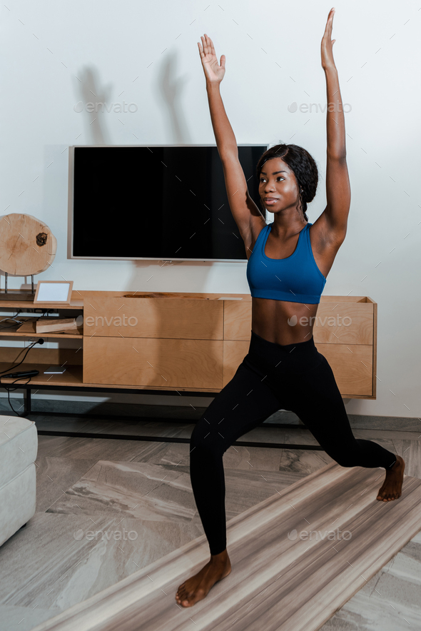 African american sportswoman standing in high lunge pose with hands in air and smiling on yoga mat