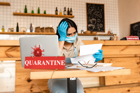 Worried cafe owner in medical mask reading document near papers, laptop and card with quarantine