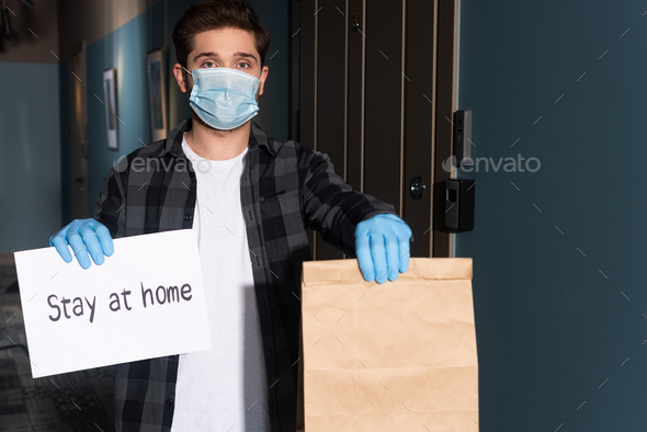 Delivery man in medical mask and latex gloves holding package and card with stay at home lettering