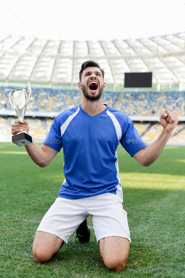 professional soccer player in blue and white uniform with sports cup standing on knees on