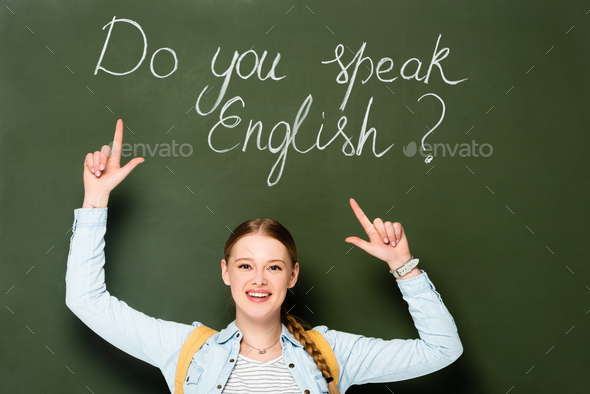 smiling girl with backpack pointing at chalkboard with do you speak English lettering