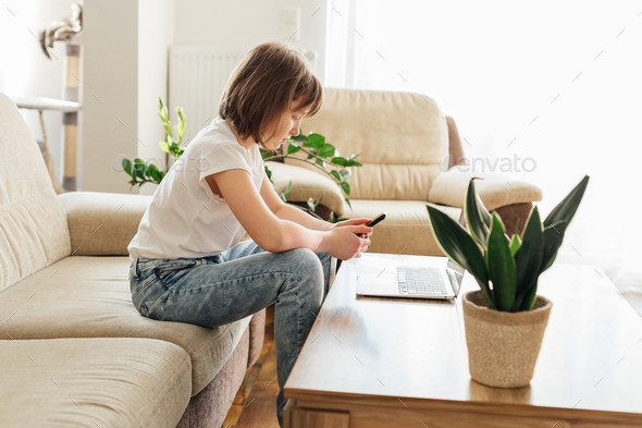 Distance learning. The girl is sitting on the couch with a phone in her hands, on the table in front - Stock Photo - Images