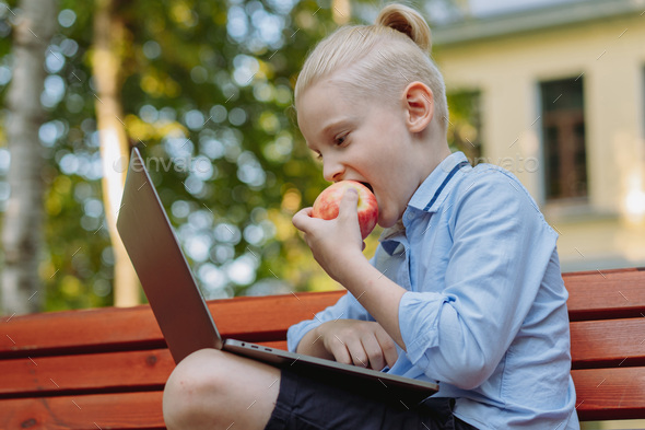 cute caucasian boy sitting on bench in park with laptop computer eating an apple - Stock Photo - Images
