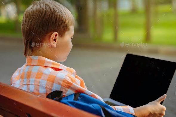 cute caucasian boy sitting on bench in park with laptop computer. Black screen - Stock Photo - Images