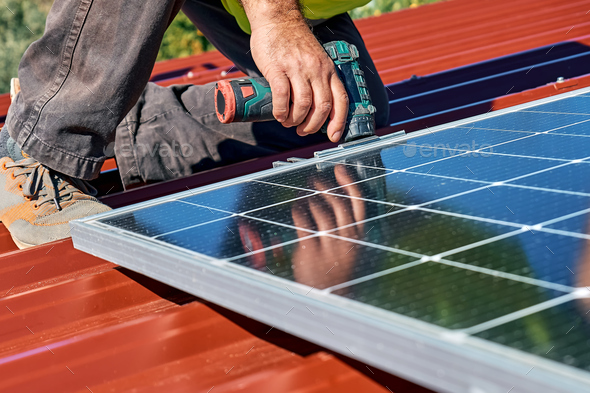 Man\'s hands with drill installing stand-alone photovoltaic solar panel system on rooftop of a house.