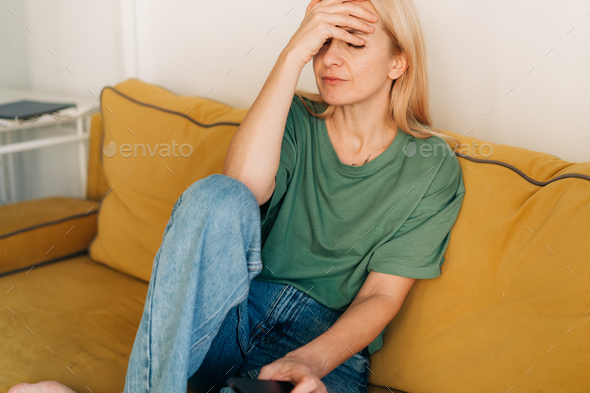 Young sad woman sitting on the couch holds her hand on her sore head. - Stock Photo - Images