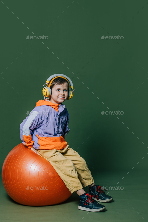 Stylish little boy in headphones sits on orange swiss ball on green background - Stock Photo - Images
