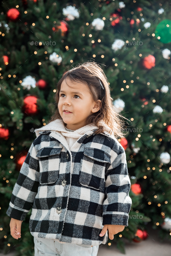 Adorable toddler girl standing next to Christmas tree outdoor, season holiday and childhood concept - Stock Photo - Images