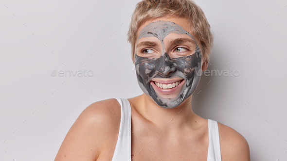Headshot of positive short haired female model applies facial clay mask for reducing fine lines and