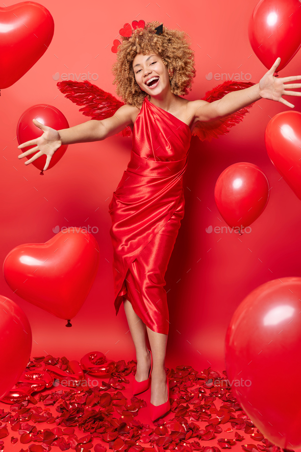 Full length shot of cheerful curly haired woman wears long dress wings and high heeled shoes going - Stock Photo - Images