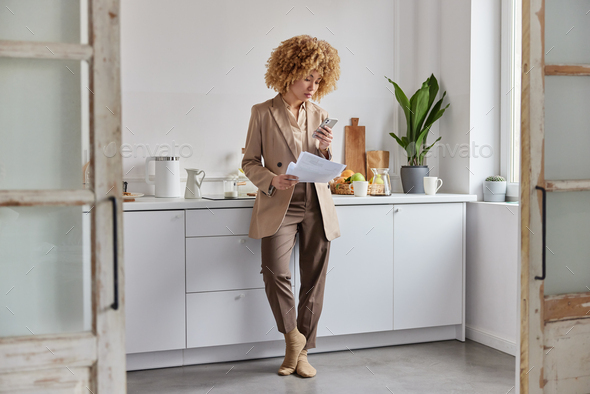 Full length shot of serious curly woman concentrated in smartphone holds paper documents uses modern - Stock Photo - Images