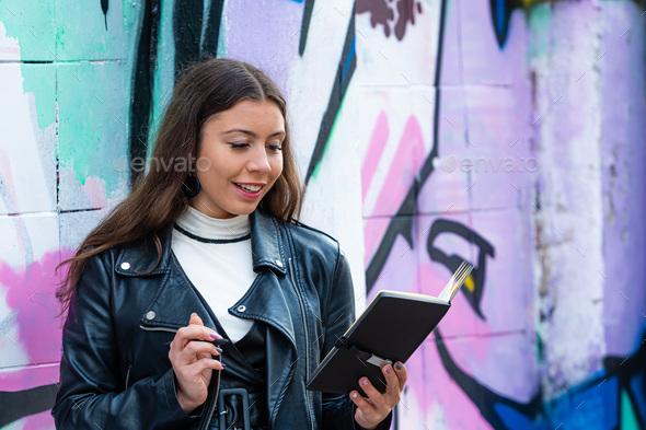 a young female leans against a wall sprayed with graffiti and makes notes in a black notebook - Stock Photo - Images
