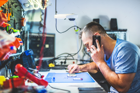 Man speaking on mobile phone repairing electronics in service shop - Stock Photo - Images