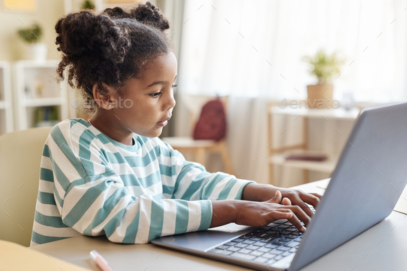 Side view cute black little girl using laptop studying online at home ...