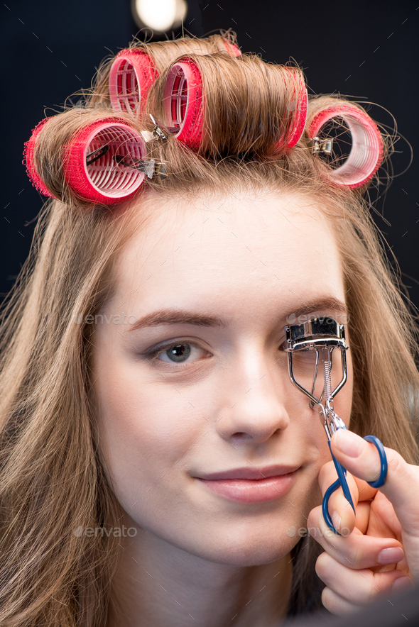 Close-up view of makeup artist correcting eyelashes with curling tongs of smiling young woman